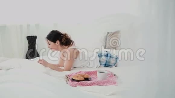 年轻女子在早餐时使用笔记本电脑躺在家里的白色床上快乐女孩吃牛角面包浏览互联网视频的预览图