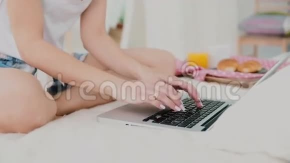 年轻女子在早餐时使用笔记本电脑坐在白色的床上近距离观看女孩聊天和喝果汁视频的预览图
