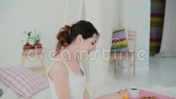 快乐的年轻女人坐在床上跳舞布鲁内特女孩使用笔记本电脑早餐时浏览互联网视频的预览图