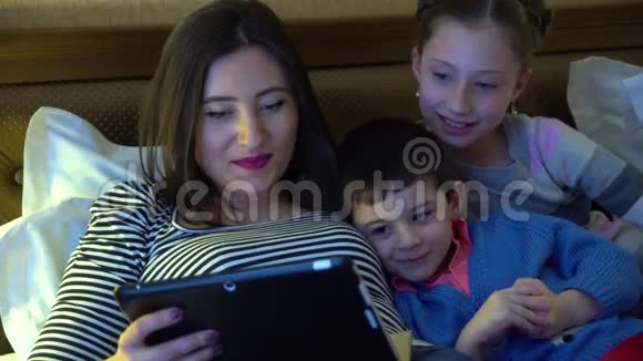 一位年轻的母亲两个孩子躺在旅馆的床上通过平板电脑使用互联网他们很开心视频的预览图