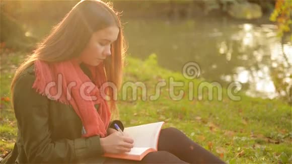 美丽的女孩坐在草地上写着一本笔记本背景是美丽的公园和湖视频的预览图