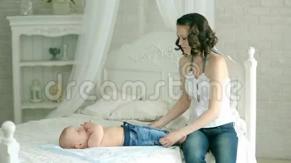 母亲给婴儿穿衣服一位年轻的母亲和一个六个月的孩子妈妈穿尿布牛仔裤和衬衫袜子6个月的孩子视频的预览图
