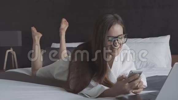 穿着白色浴袍和戴着智能手机和笔记本电脑的眼镜的女人躺在床上微笑在线商务视频的预览图