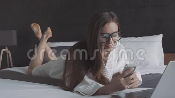 穿着白色浴袍和戴着智能手机和笔记本电脑的眼镜的女人躺在床上在线商务视频的预览图
