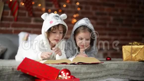 两个可爱的小兄弟姐妹在圣诞树附近的床上看书里面有灯光和照明两个幸福的家庭视频的预览图