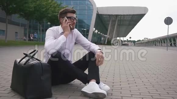 在机场戴眼镜的英俊商人拿着行李坐在地上通过电话交谈试图解决这个问题视频的预览图