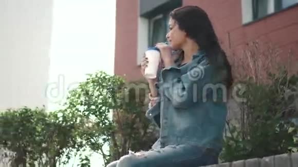 穿着牛仔夹克在街上喝着纸杯咖啡的年轻漂亮女孩视频的预览图