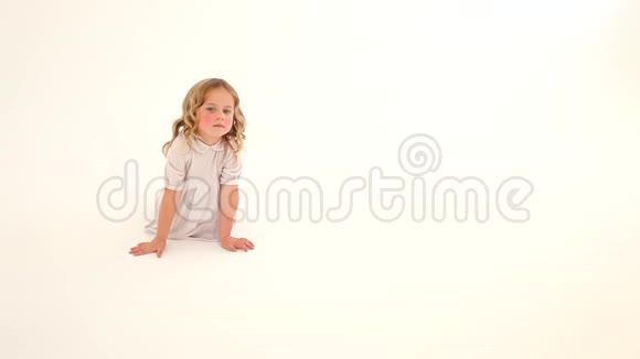 幼儿小女孩红润的脸颊金色的卷发白色的连衣裙坐在地上手指白色的背景视频的预览图