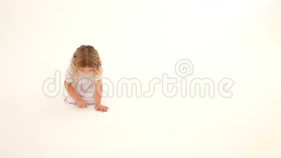 婴儿学步的孩子女孩金发卷发红润的脸颊白色的连衣裙坐着抽着心手指在地板上白色的背景视频的预览图