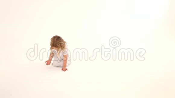 婴儿学步的孩子女孩红润的脸颊金色的卷发白色的连衣裙坐着抽着心手指在地板上白色的背景视频的预览图