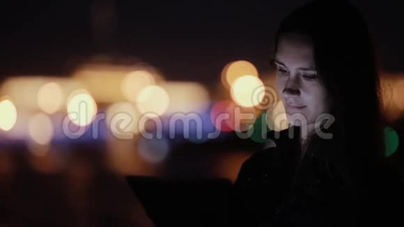 美丽的年轻女人用平板电脑晚上微笑背景上模糊的彩色城市灯光现代技术视频的预览图