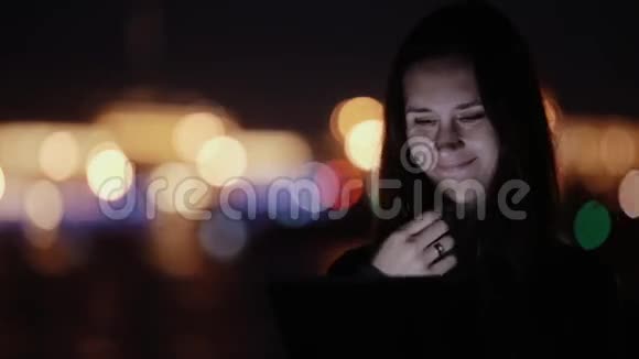 年轻漂亮的女人用平板电脑晚上微笑背景上模糊的彩色城市灯光现代技术视频的预览图