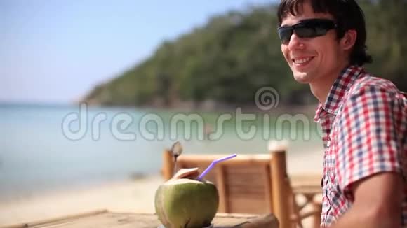 英俊的年轻快乐男子戴着太阳镜在海滩咖啡馆喝着新鲜的椰子汁在棕榈树上欣赏海景视频的预览图