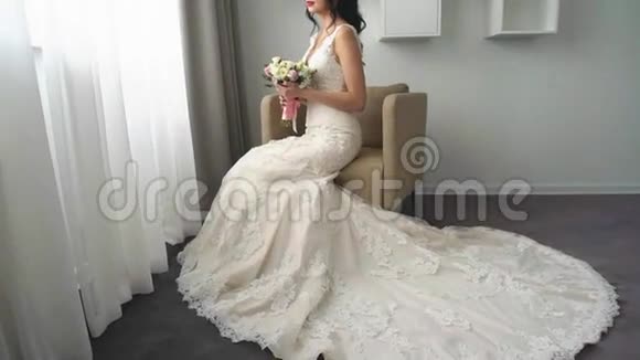 迷人的年轻新娘坐在扶手椅上穿着婚纱手里捧着一大束鲜花做着白日梦视频的预览图