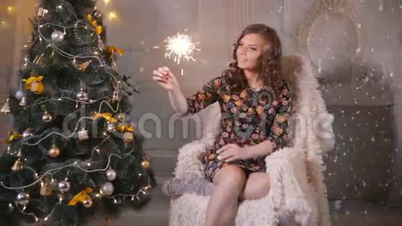 迷人的年轻女子在靠近圣诞树的椅子上举着火花孟加拉火庆祝圣诞节视频的预览图