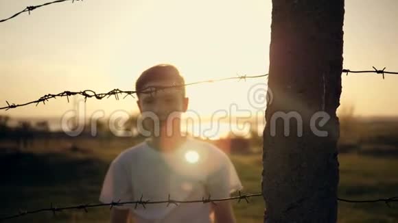那个男孩在铁丝网后面日落时手拿着电线移民概念一个孩子的剪影视频的预览图