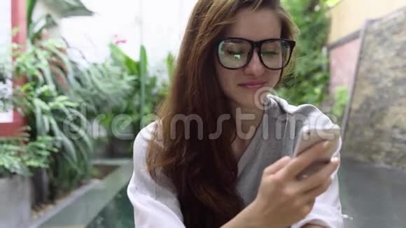 戴眼镜的女人在热带国家游泳池附近使用智能手机视频的预览图