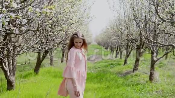一个穿着粉红色裙子的金发女人走在盛开的花园旁那里有树视频的预览图
