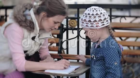 小漂亮女孩和她的母亲坐在公园的长凳上手里拿着她的笔记本她穿着一件外套她是视频的预览图