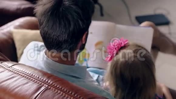 上景年轻的父亲和他的金发小女儿正在餐厅的皮革沙发上读一个故事4K视频的预览图