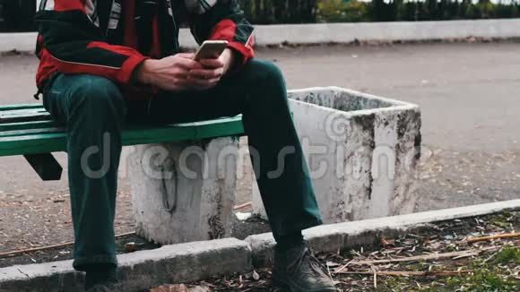 年轻人正坐在公园的绿色长凳上等着别人在智能手机上打字视频的预览图