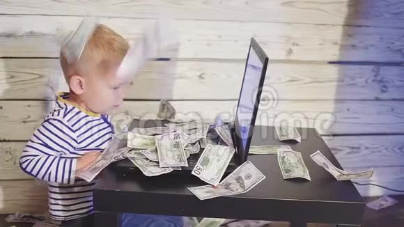 两岁的男孩有一台电脑钱也在下降微笑的男孩坐在桌子上手提电脑和空中的美元钞票视频的预览图