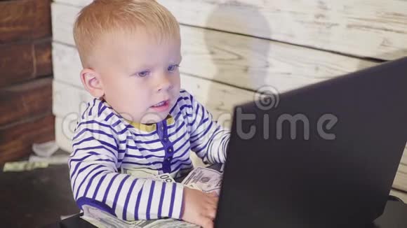 两岁的男孩有一台电脑钱也在下降微笑的男孩坐在桌子上手提电脑和空中的美元钞票视频的预览图