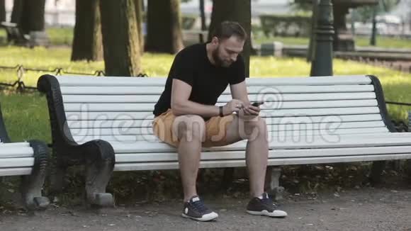 有魅力的年轻人坐在公园的长凳上用智能手机发送短信视频的预览图