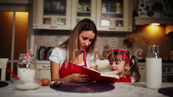穿着红色围裙的妈妈和女儿在厨房里读着笔记本上的食谱蛋糕视频的预览图