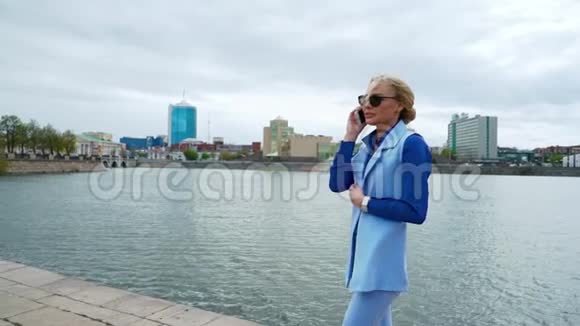 年轻的金发美女在河边的公园里散步在手机上聊天蓝色衬衫商务套装视频的预览图