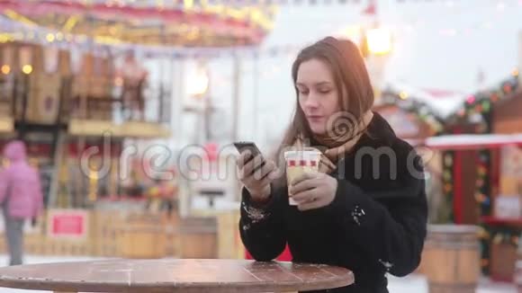 美丽的年轻女子在旋转木马前的圣诞集市上使用智能手机喝着热茶视频的预览图