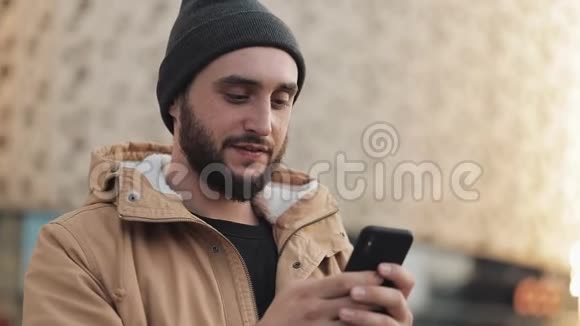 快乐的年轻胡须男在购物中心附近的街道上使用智能手机他穿着一件秋装和一顶针织帽子视频的预览图