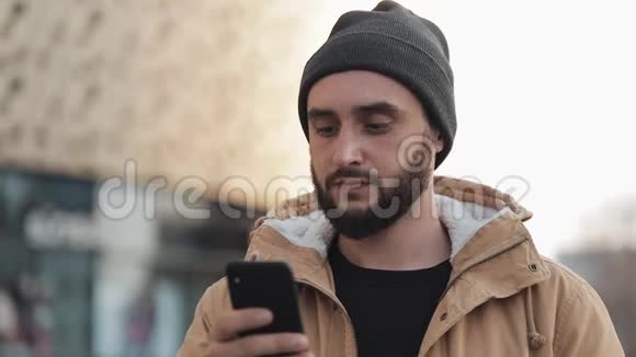 快乐的年轻胡须男在购物中心附近的街道上使用智能手机他穿着一件秋装和一顶针织帽子视频的预览图