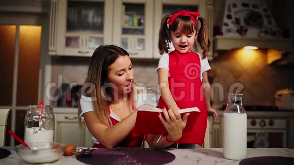 穿着红色围裙的妈妈和女儿在厨房里读着笔记本上的食谱蛋糕视频的预览图