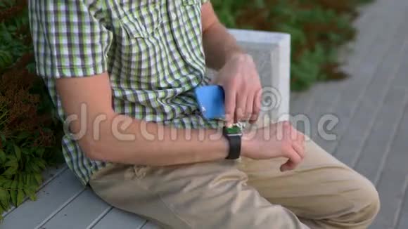 穿着休闲服的男人用他的小玩意智能手表和智能手机坐在长凳上视频的预览图