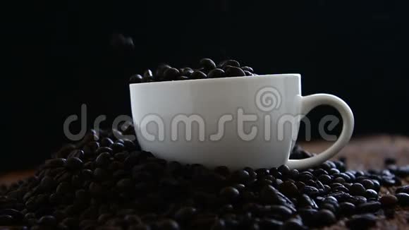慢动作的烤咖啡豆掉进了一堆咖啡豆和木地板上的白色陶瓷咖啡杯里视频的预览图