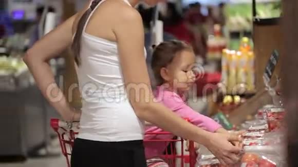 年轻的母亲带着小女儿在手推车里挑选超市里的西红柿视频的预览图