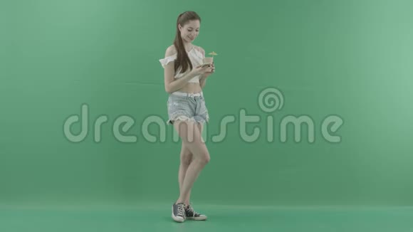 年轻漂亮的女人在绿色背景下喝鸡尾酒时在智能手机上发短信视频的预览图