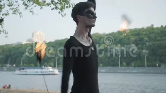 年轻的苗条男子穿着黑色的衣服和面具表演一场表演与火焰站在河岸有技巧的消防艺术家视频的预览图
