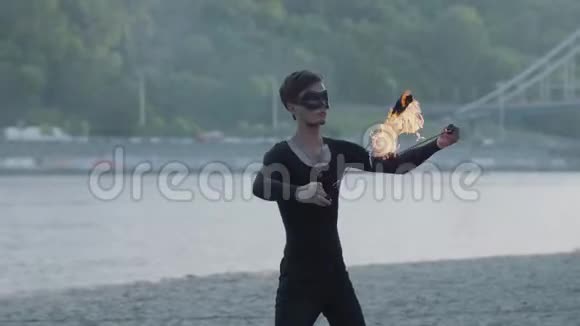 年轻的苗条男子穿着黑色的衣服和面具表演一场表演与火焰站在河岸有技巧的消防艺术家视频的预览图