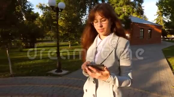 漂亮的女商人戴着眼镜走在公园的小巷里手里拿着平板电脑一个生意清淡的女孩视频的预览图