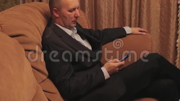严肃的商人坐在沙发上用智能手机看信息视频的预览图