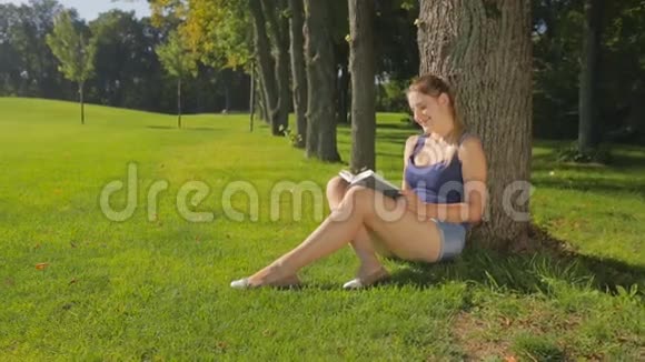 美丽的年轻女子坐在公园的树下看书视频的预览图