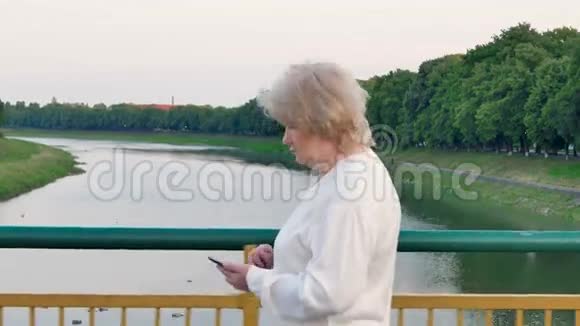 一位老年妇女在河上的桥上行走使用智能手机视频的预览图