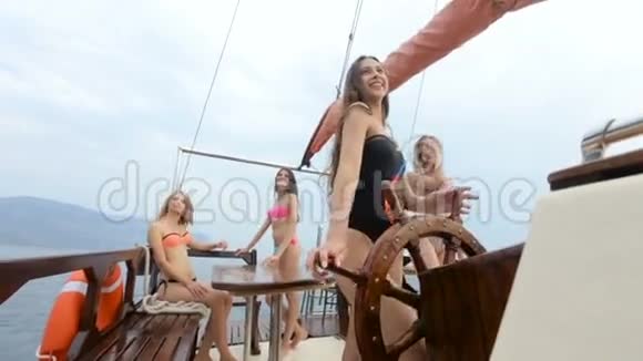穿着泳衣的漂亮女孩站在游艇轮后带着红色的帆而她的朋友们在玩着视频的预览图