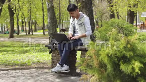 亚洲男人坐在公园里用笔记本电脑工作视频的预览图