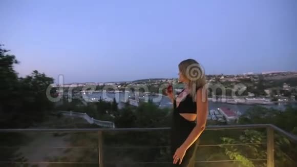 迷人的年轻金发碧眼身穿黑色连衣裙站在夏季露台上欣赏城市美景喝着香槟视频的预览图