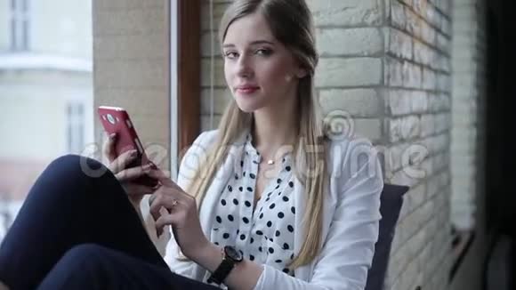 年轻漂亮的金发女郎坐在现代的办公室里用手机发短信然后看着相机视频的预览图