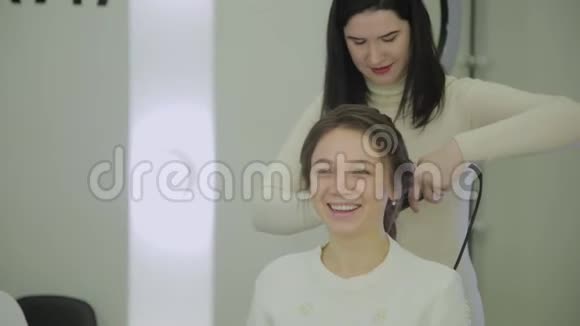 一位棕色头发的美女正在接受理发师用卷发熨斗治疗实时手持设备视频的预览图