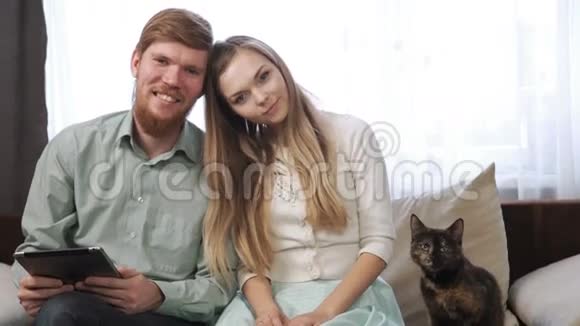 一对夫妇一个年轻的男人和一个女人他们的猫坐在家里的沙发上看着平板电脑微笑着视频的预览图